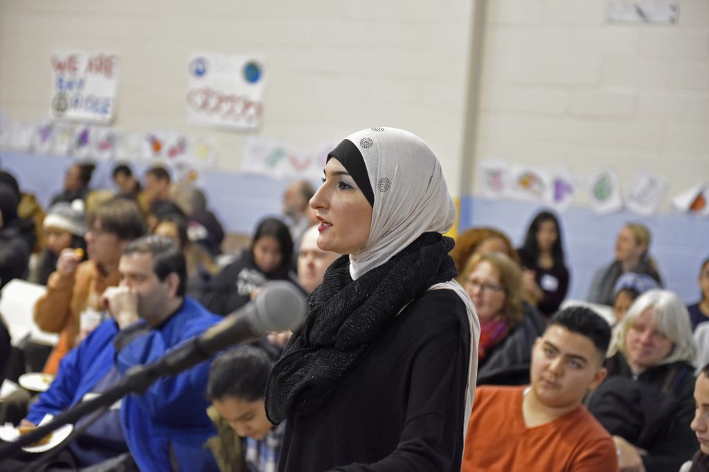 woman in a headwrapping speaking in front of a crowd in a school about a stabbing in boston